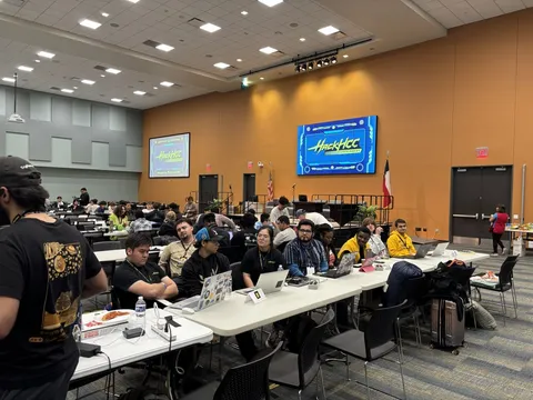 Hackers working at long tables in the HackHCC main hall, event branding on the screens behind them
